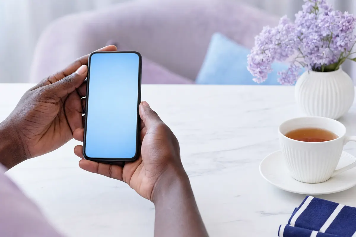 Hands holding a smartphone with blank blue screen beside a cup of tea